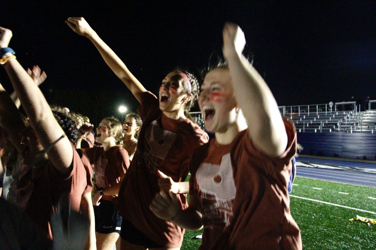 Arden Village Powderpuff players jump up and down while cheering for their teammates. The players each donned their hall t-shirts and red face paint, some opting to wear cleats and football gloves.