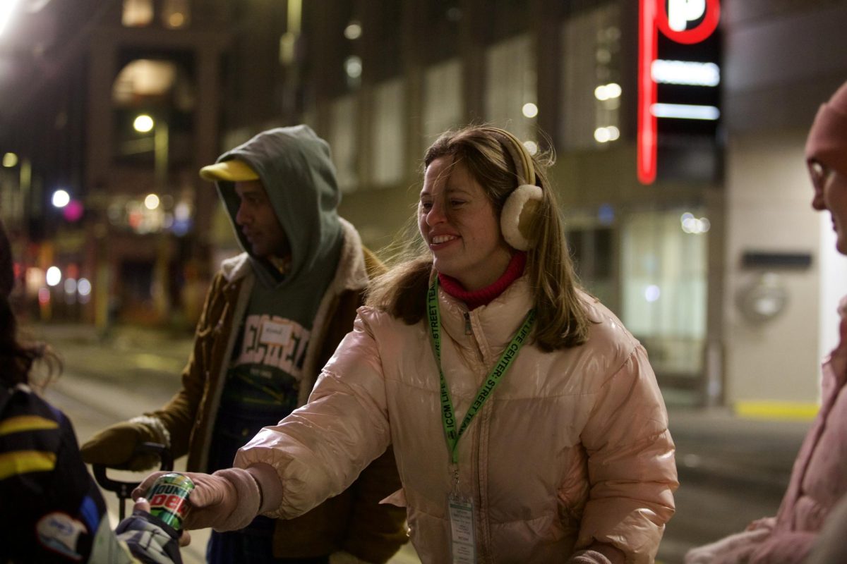 Street Team volunteer captain Sarah Engwall hands a Mountain Dew to a woman waiting for the light-rail Friday, Feb. 27, 2026 in Minneapolis. Engwall said volunteering for the ICCM Life Center’s Friday Night Street Team has challenged her faith. “[It has] been so humbling when meeting people who were in great spots but lost everything,” Engwall said. “They still had their faith.” (Photo by Kathryn Kovalenko)
