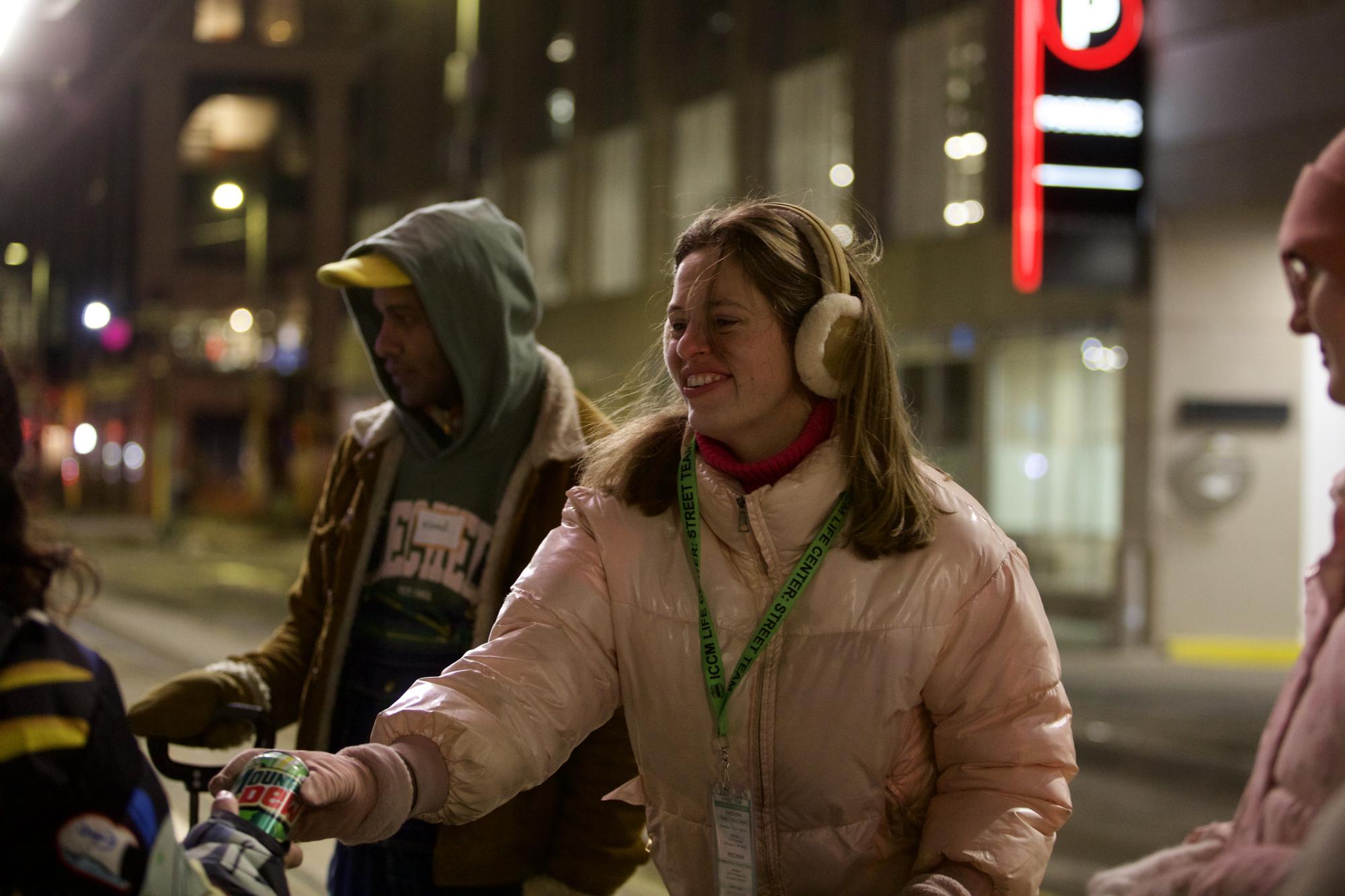 Street Team volunteer captain Sarah Engwall hands a Mountain Dew to a woman waiting for the light-rail Friday, Feb. 27, 2026 in Minneapolis. Engwall said volunteering for the ICCM Life Center’s Friday Night Street Team has challenged her faith. “[It has] been so humbling when meeting people who were in great spots but lost everything,” Engwall said. “They still had their faith.” (Photo by Kathryn Kovalenko)
